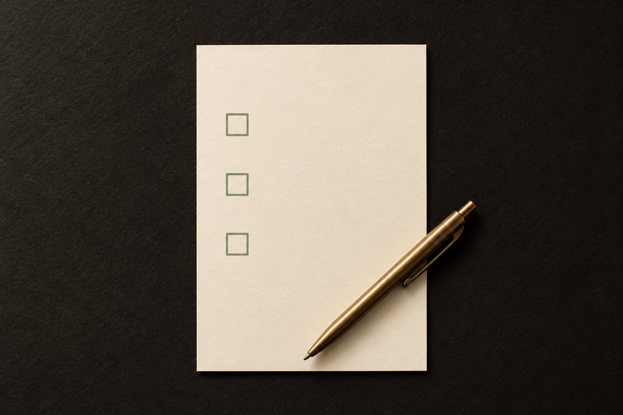 A cream card with three empty sage-green checkbox squares lies beneath a diagonally placed brass pen.